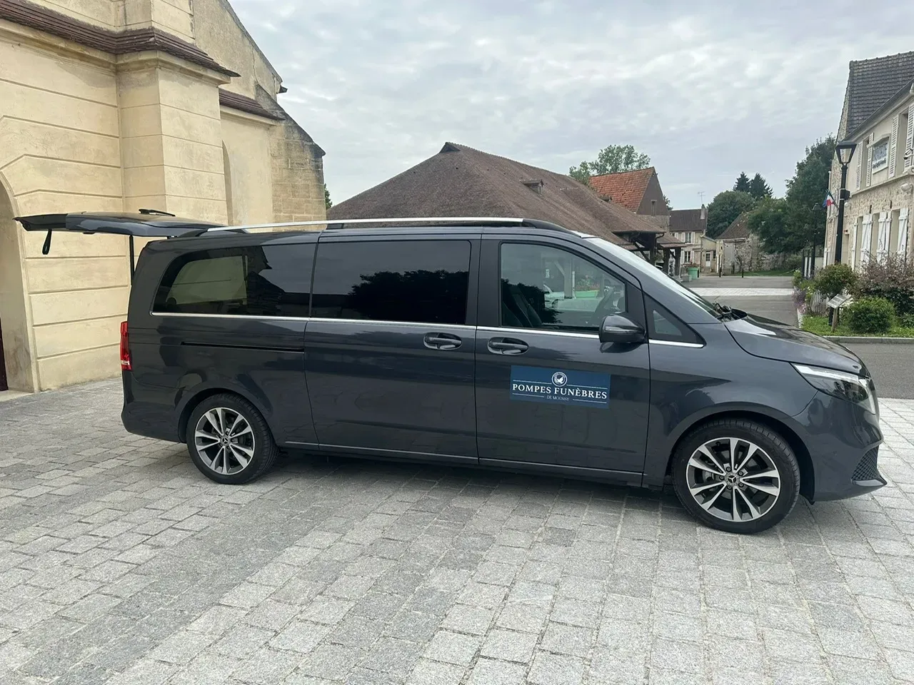 Véhicule de pompes funèbres gris foncé stationné devant un bâtiment en pierre.