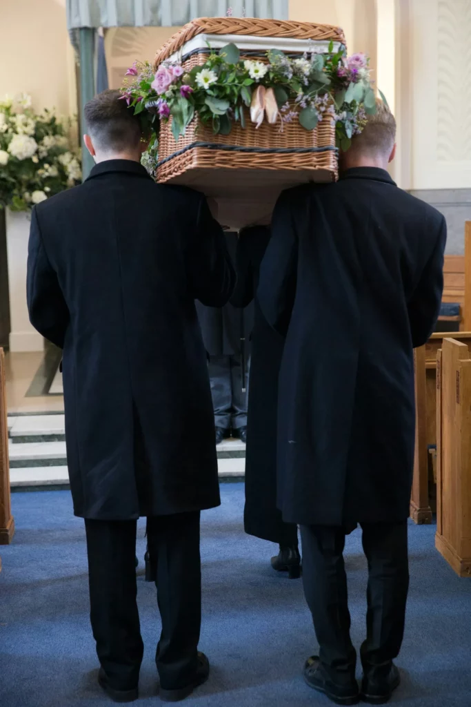 Porteurs funéraires en costume sombre portant un cercueil en osier fleurie dans une église.